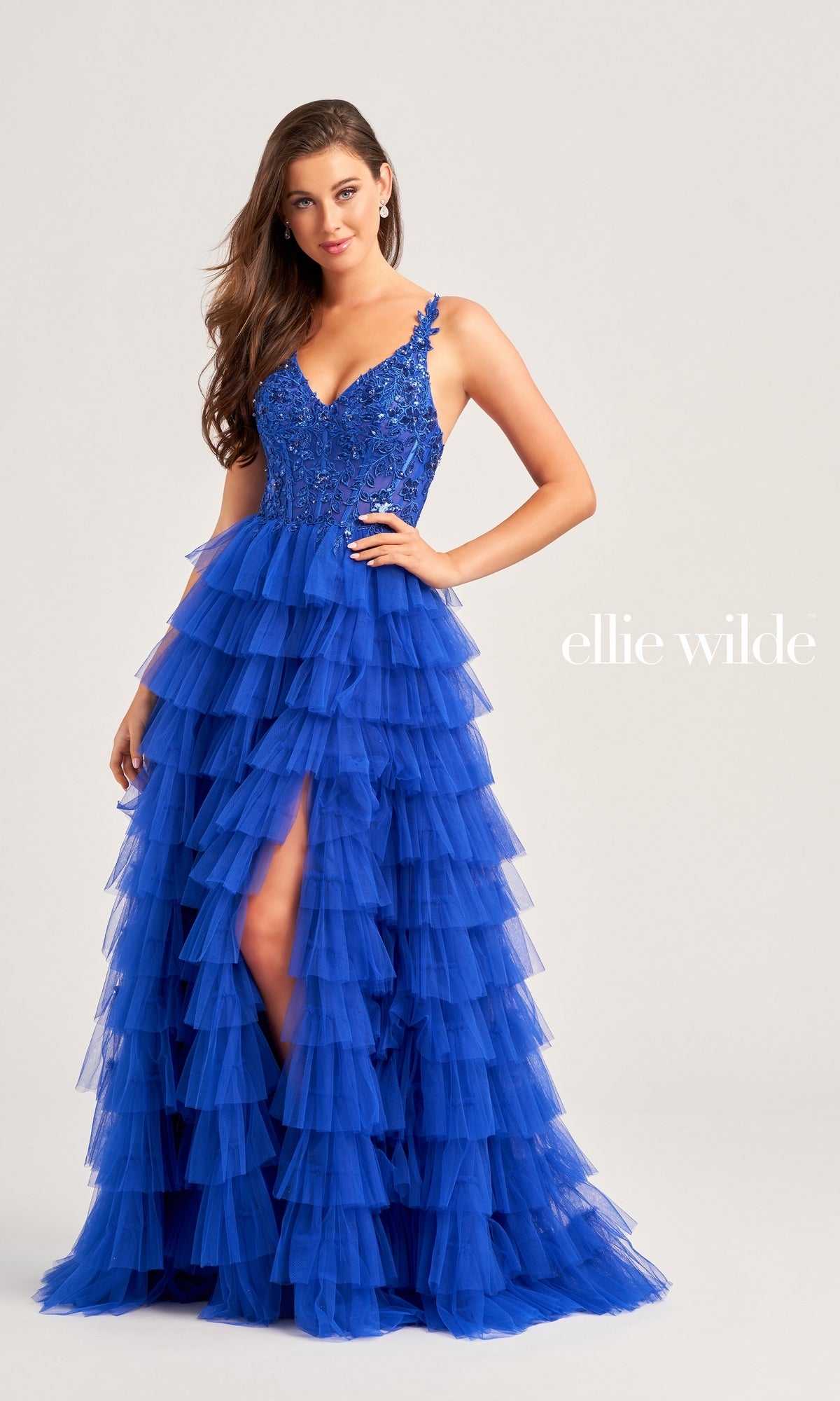 A woman wears the Tiered Ellie Wilde EW35059 Designer Ball Gown, featuring a sequin corset bodice and ruffled tiered skirt, posing against a light background with 