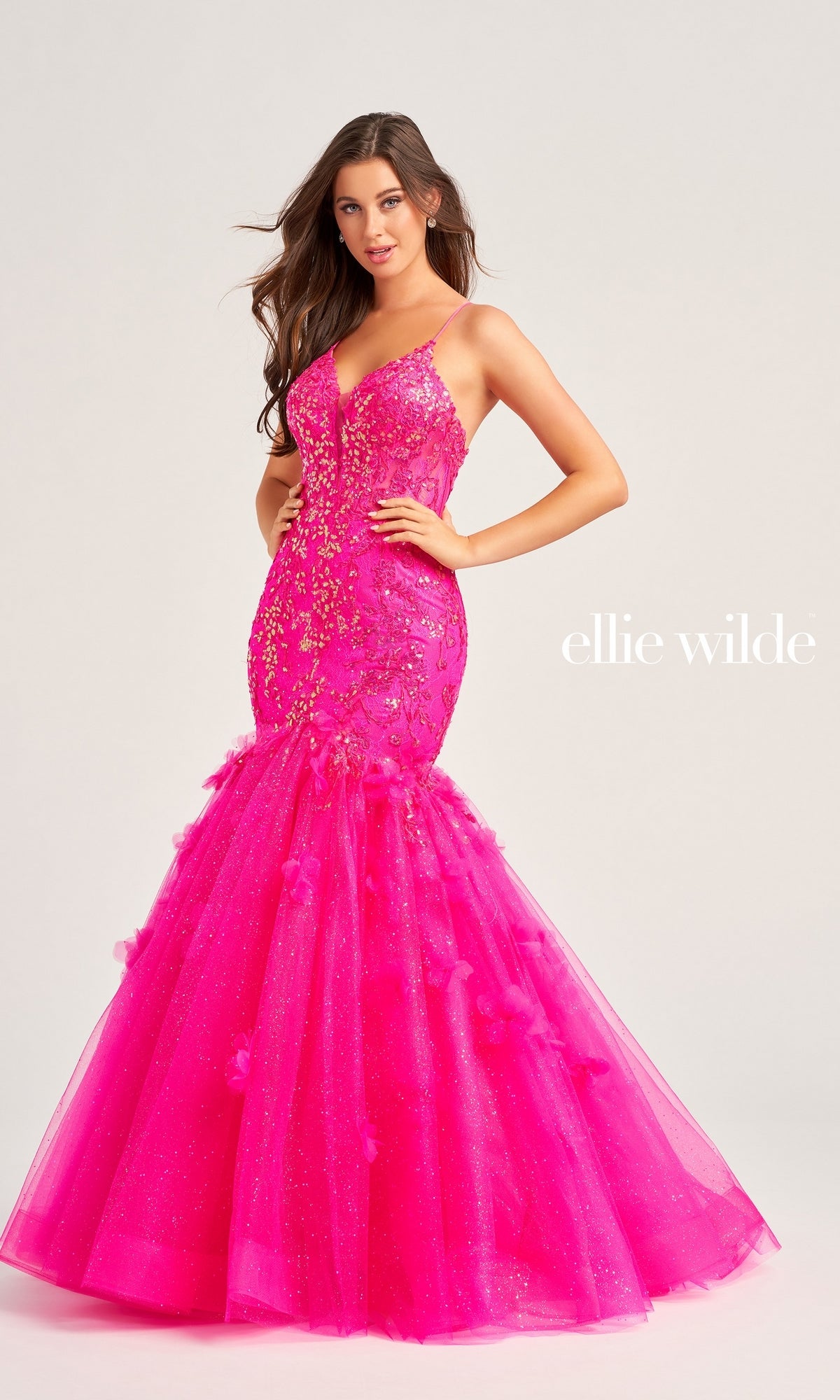 A woman models the Ellie Wilde Designer Mermaid Prom Dress EW35080 in vibrant pink with floral and sparkly embellishments, standing against a plain background. 