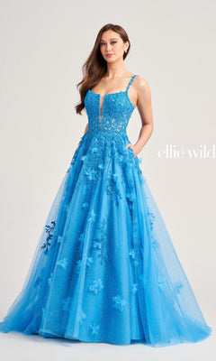 A woman wearing the Glitter-Tulle Ellie Wilde Long Prom Gown EW35081, featuring a sheer bodice, floral appliques, and a tulle skirt, stands against a plain background.