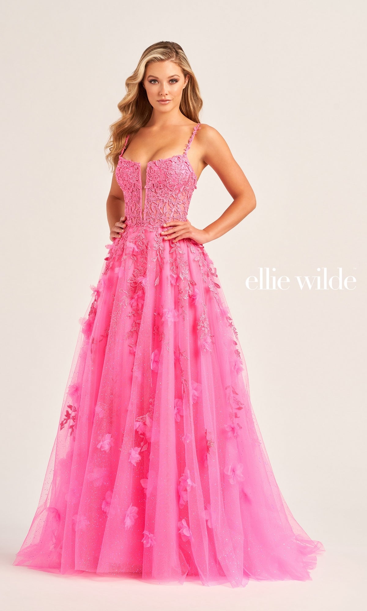 A woman in the Glitter-Tulle Ellie Wilde Long Prom Gown EW35081, a bright pink floor-length dress with floral appliques and thin straps, stands against a plain background. 