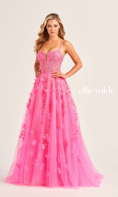 A woman in the Glitter-Tulle Ellie Wilde Long Prom Gown EW35081, a bright pink floor-length dress with floral appliques and thin straps, stands against a plain background. "Ellie Wilde" appears beside her.