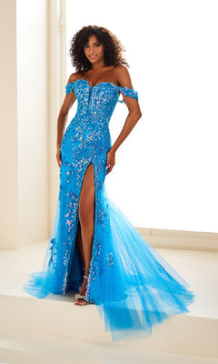 A woman smiles at the camera indoors, wearing the Off-Shoulder Ellie Wilde Designer Dress EW35082—a bright blue evening gown with sequin embellishments, a front slit, and tulle train.