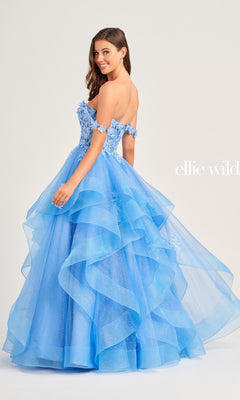 A woman stands smiling over her shoulder in the Ellie Wilde Long Designer Prom Ball Gown EW35084, a strapless, tiered blue glitter tulle dress featuring floral appliqué details.