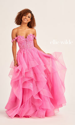 A woman stands against a plain background with "ellie wilde" text, wearing the Ellie Wilde Long Designer Prom Ball Gown EW35084—an off-the-shoulder pink dress featuring ruffles, floral embellishments, and glitter tulle.