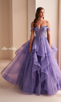A woman stands indoors wearing the Ellie Wilde Long Designer Prom Ball Gown EW35084, a strapless lavender evening gown with off-the-shoulder sleeves, floral appliques, tiered layers, and glitter tulle.