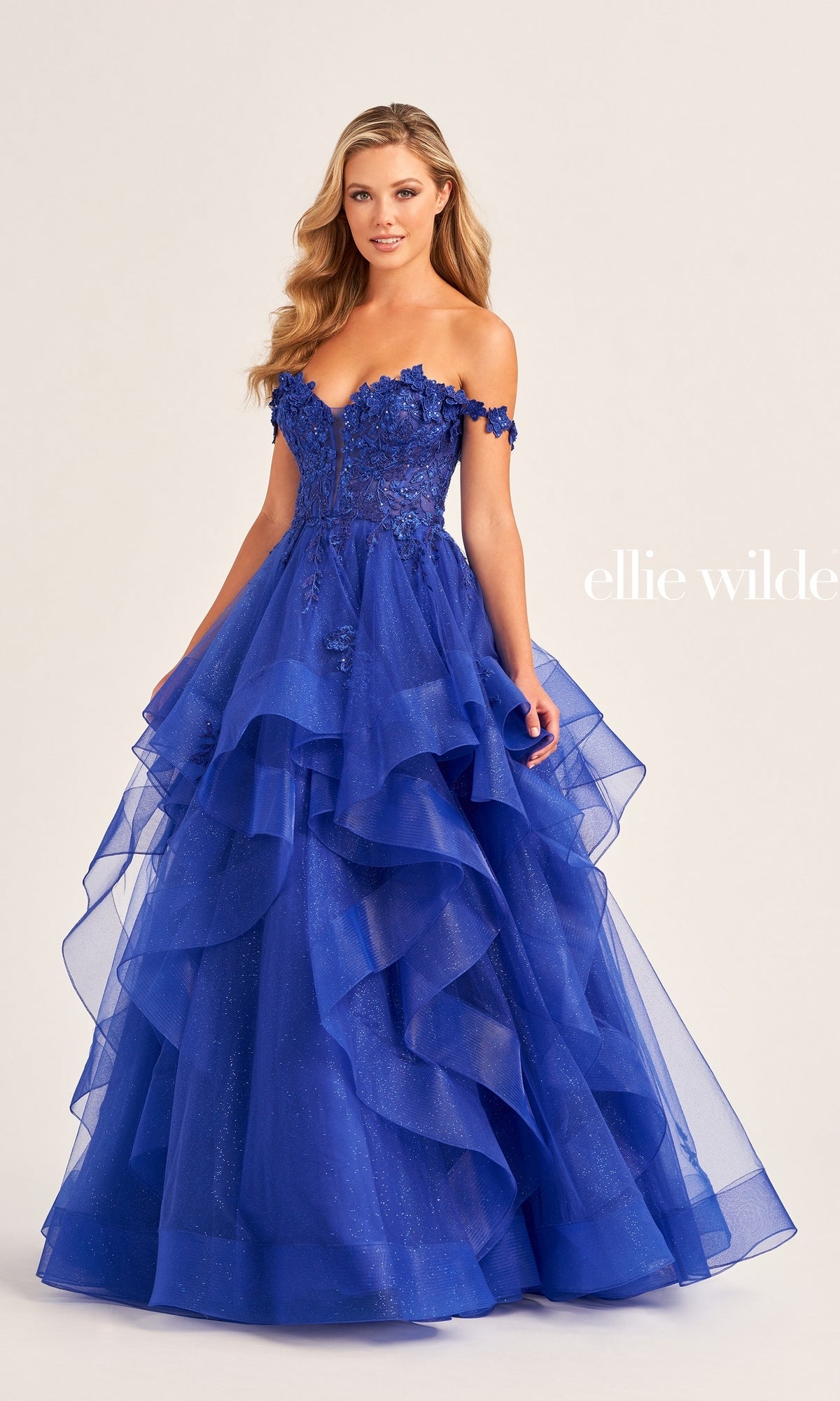 A woman wears the Ellie Wilde Long Designer Prom Ball Gown EW35084, a bright blue off-the-shoulder dress with floral appliqués, a fitted bodice, and layered glitter tulle, standing against a plain background.