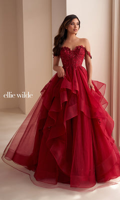 A woman wears the Ellie Wilde Long Designer Prom Ball Gown EW35084— a strapless, layered deep red glitter tulle dress with floral accents and off-the-shoulder sleeves. She stands by an indoor window in this stunning designer gown.