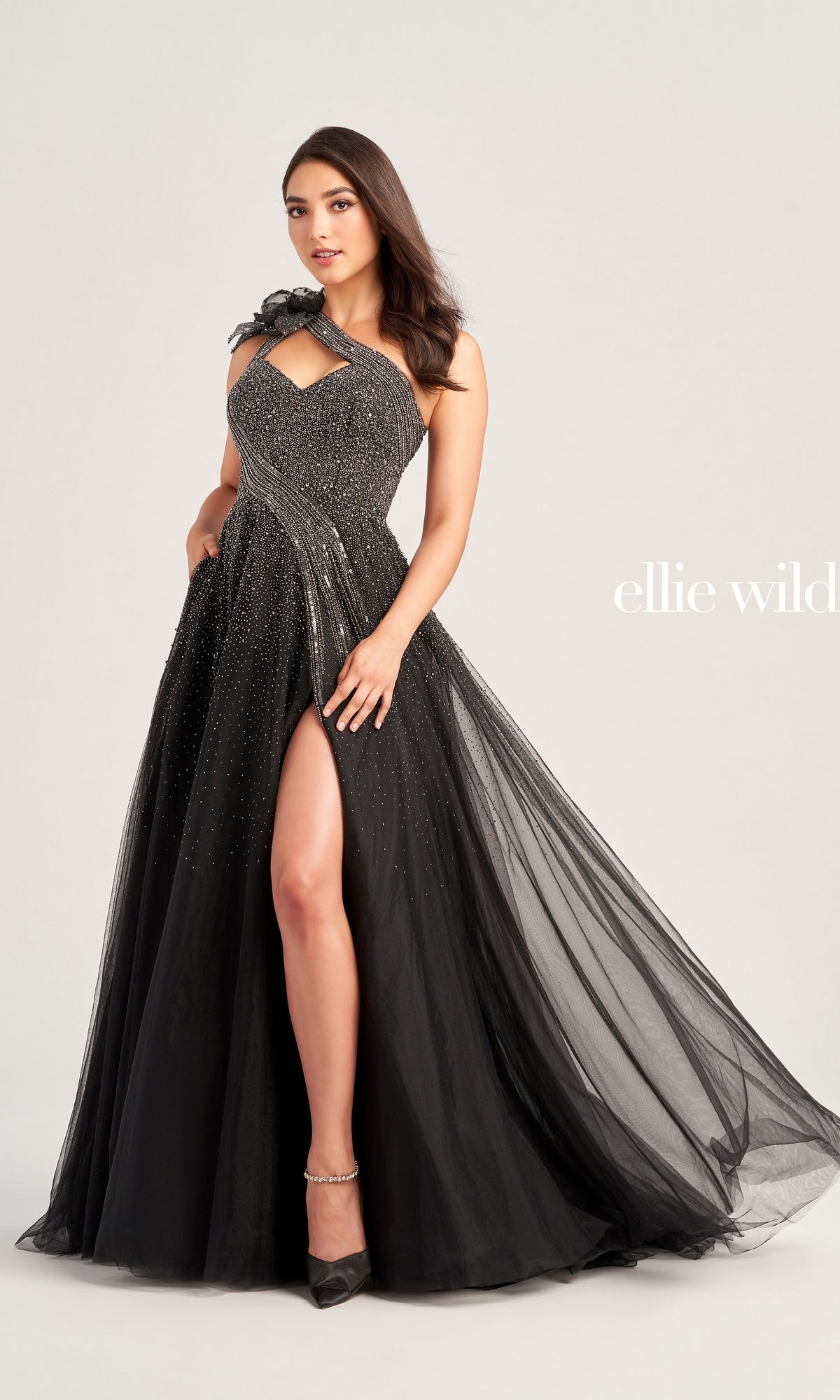 A woman wears the Long Prom Dress EW35086 by Ellie Wilde, a black one-shoulder gown with a high slit, sparkling embellishments, and floral shoulder detail, standing against a plain background.