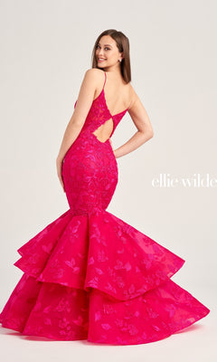 A woman wears the Ellie Wilde Long Lace Mermaid Prom Dress EW35092 in bright pink, featuring lace details, a layered skirt, and an open back, standing against a plain light background.