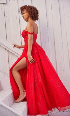 A woman in the Ellie Wilde Off-Shoulder Long Prom Dress EW35101, a bright red gown with a thigh-high slit, stands on white stairs against a white wooden wall, looking over her shoulder.