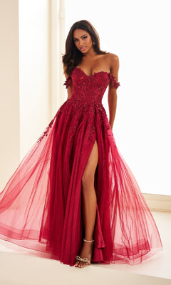 Woman wearing the Ellie Wilde Off-Shoulder Long Prom Dress EW35101, a floor-length red glitter tulle gown with floral embellishments and a side slit, paired with high-heeled sandals, standing by a window indoors.