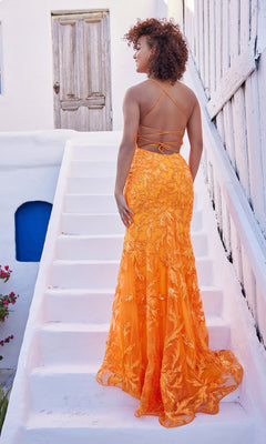 A woman stands on white steps outdoors, facing away, wearing the Ellie Wilde Long Designer Formal Dress EW35109—a backless, floor-length gown with embroidered lace in a striking orange hue.