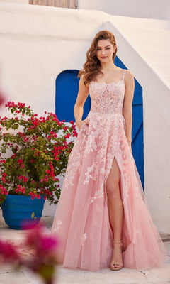 A woman stands outdoors in front of a white building with a blue door, wearing the Glitter-Tulle Ellie Wilde Long Prom Dress EW35114—a pink, floral-embroidered gown featuring a high slit and spaghetti straps.