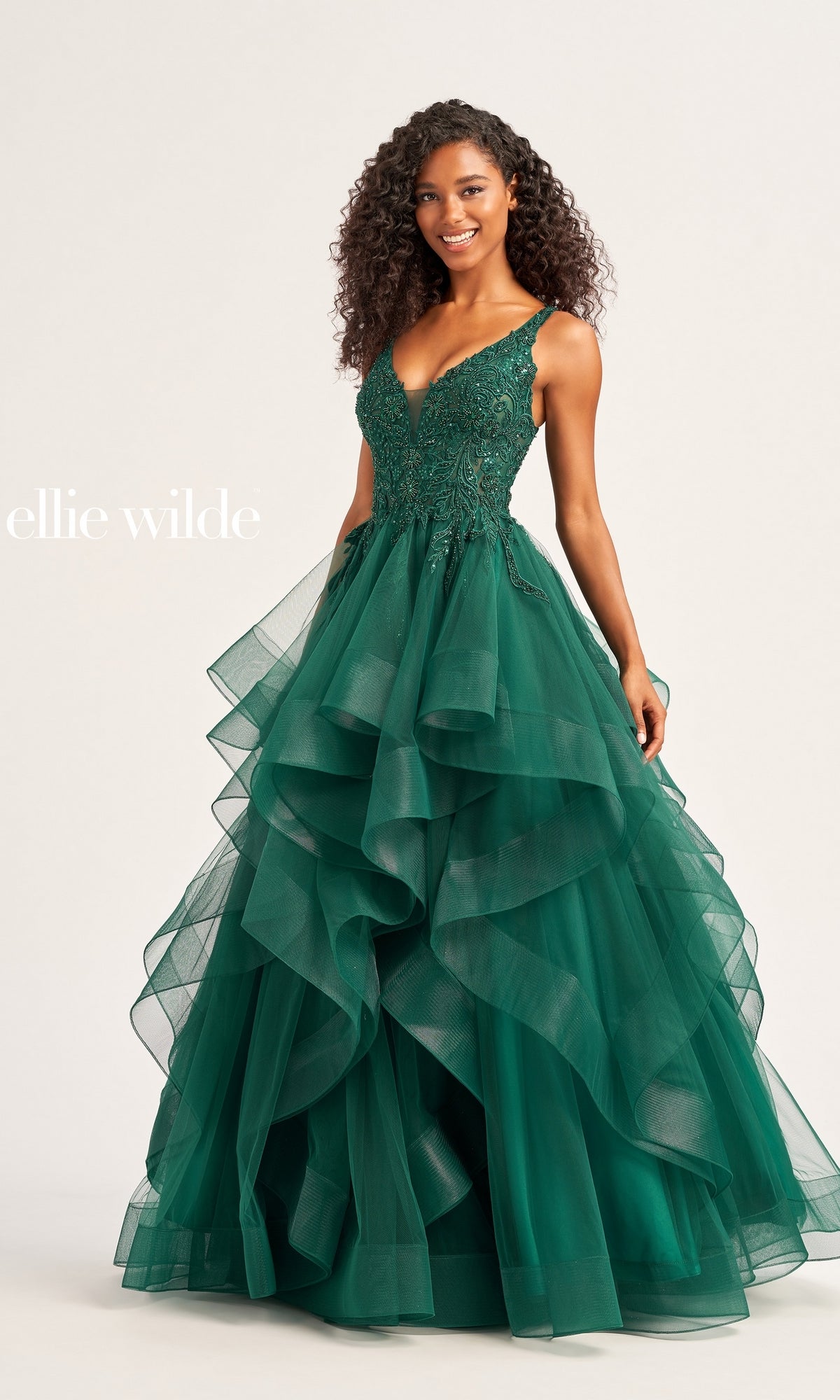 A woman stands against a plain background wearing the Ellie Wilde Tiered Long A-Line Prom Dress EW35119, featuring a sleeveless emerald green lace bodice with beaded details and a layered tulle skirt. 