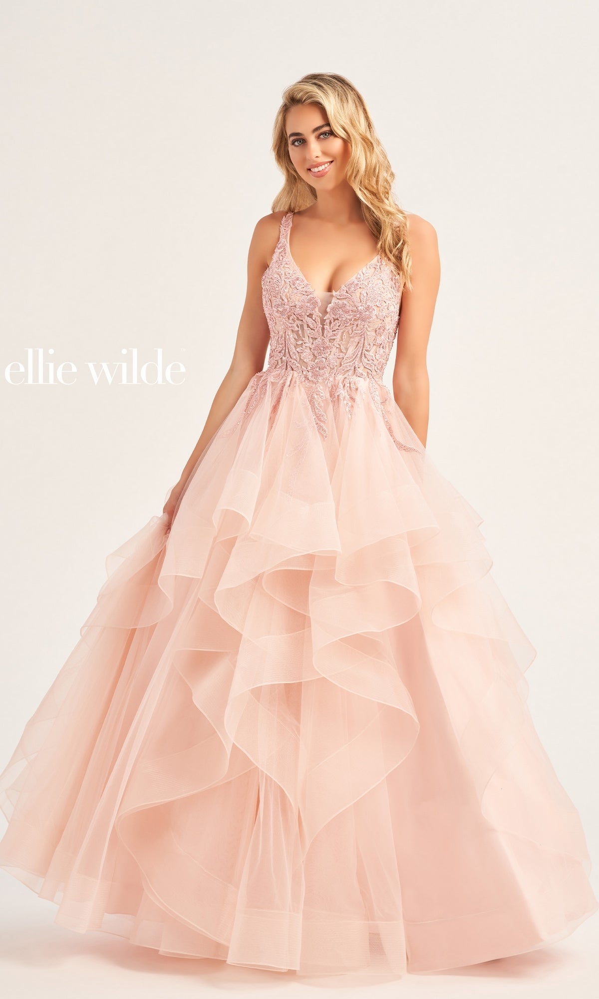 A woman in a light pink, sleeveless Ellie Wilde Tiered Long A-Line Prom Dress EW35119 with a lace bodice stands against a plain background. 