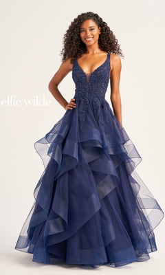 A woman smiles against a plain light background, wearing the sleeveless Ellie Wilde Tiered Long A-Line Prom Dress EW35119 in deep blue with a beaded bodice. The text "ellie wilde" is displayed on the left.
