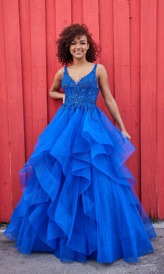 A woman in a vibrant blue Ellie Wilde Tiered Long A-Line Prom Dress EW35119 poses smiling in front of a red wooden wall.