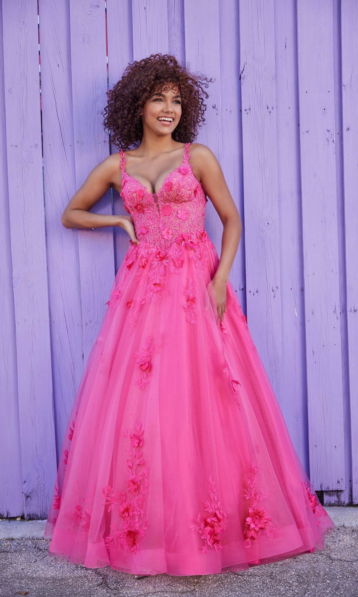 A woman smiles against a purple wooden fence wearing the Ellie Wilde Bright Pink Prom Ball Gown EW35122 featuring a corset-style bodice.