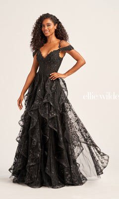 A woman wears the Ellie Wilde Ruffled Long Prom Ball Gown EW35218, a black, off-the-shoulder dress featuring ruffles, glittery details, and beaded lace appliques, while standing against a plain background.