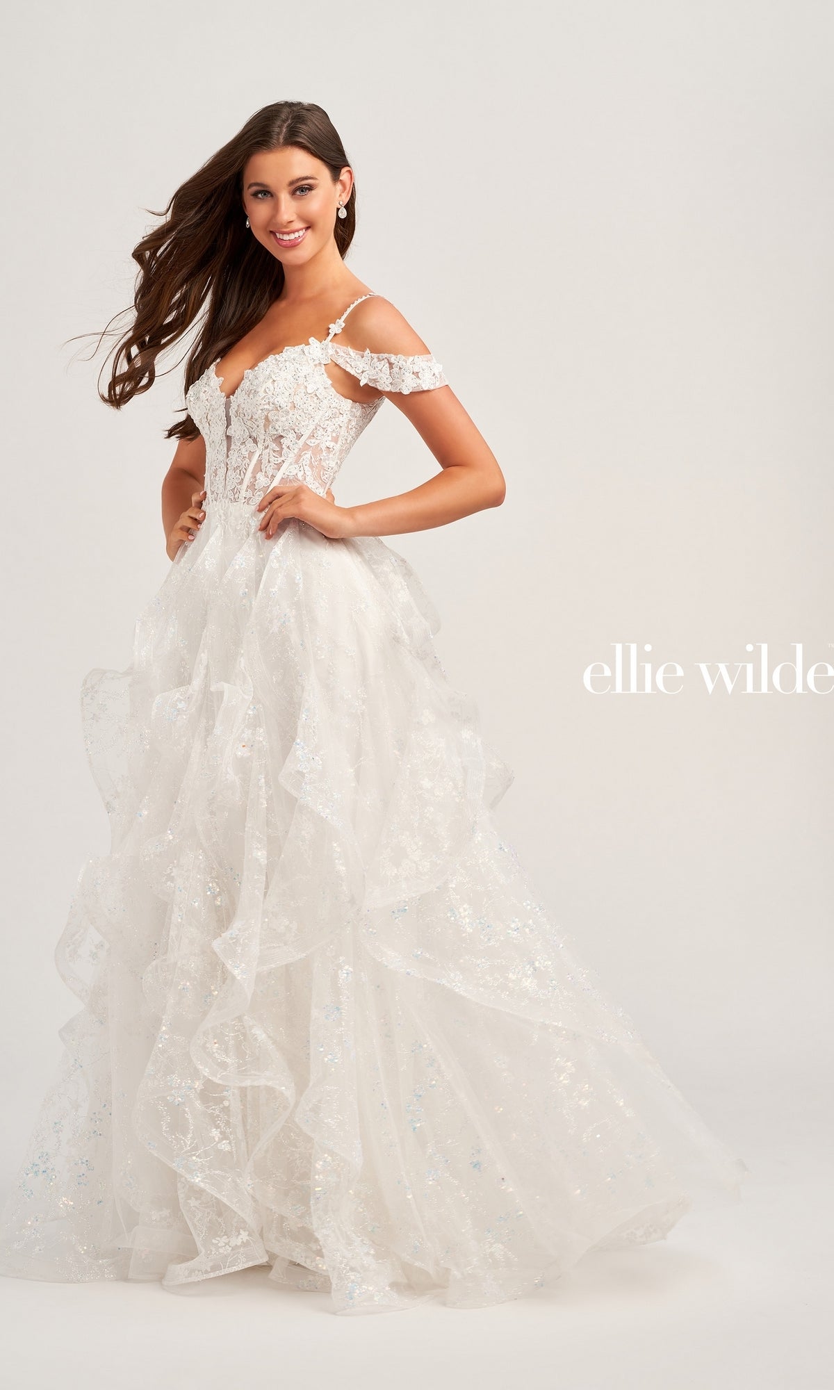 A woman smiles in the Ellie Wilde Ruffled Long Prom Ball Gown EW35218, a white off-the-shoulder dress with ruffles, beaded lace appliques, and sequin details; 