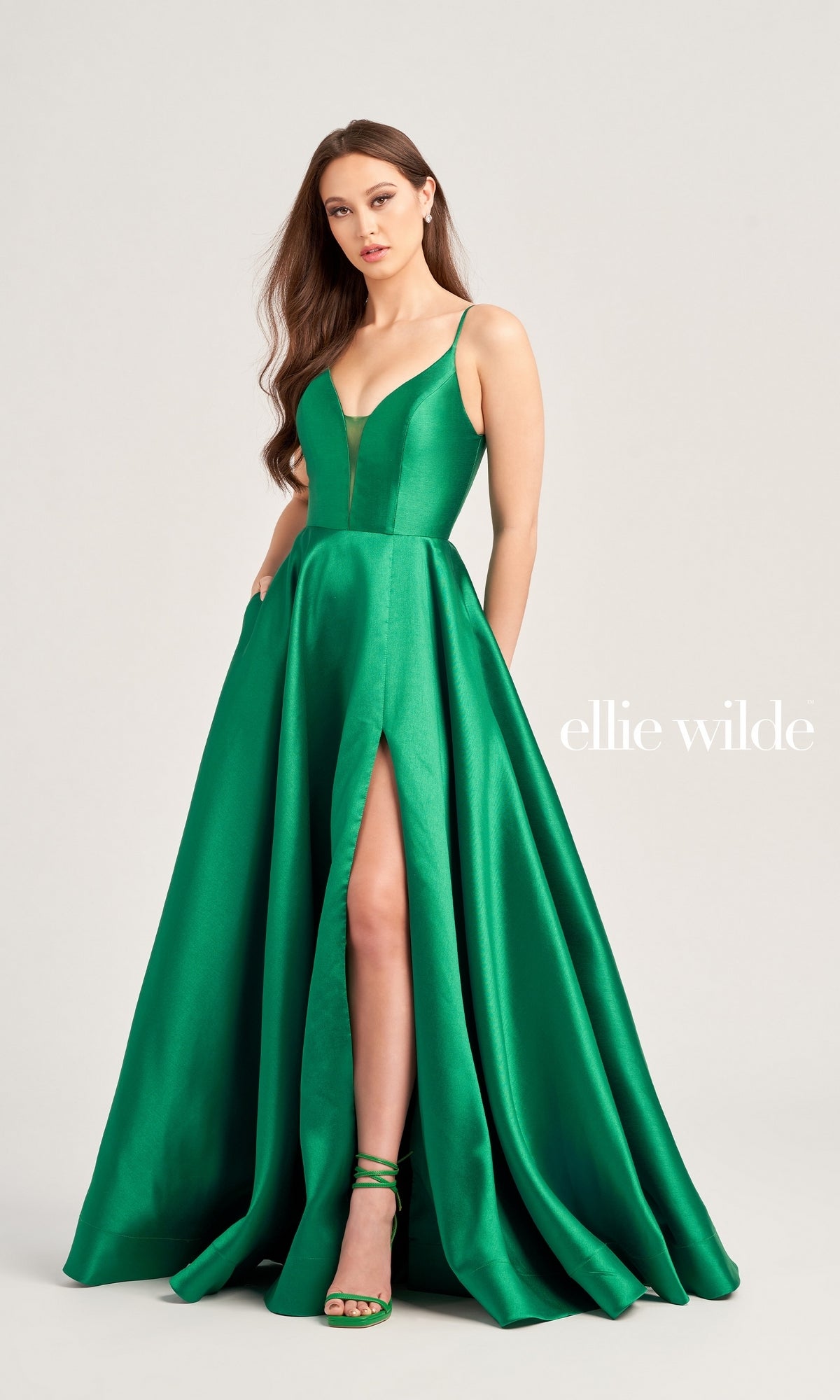 A woman wears the Ellie Wilde Long A-Line Satin Prom Dress EW35232 in emerald green, featuring a sleeveless design, floor-length satin skirt with a deep neckline and high slit, paired with matching green heels. 
