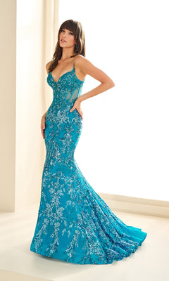 A woman in the Ellie Wilde EW36003 Sequin-Print Mermaid Prom Dress stands by a sunlit indoor window; the fitted, floor-length turquoise gown features intricate silver embroidery.