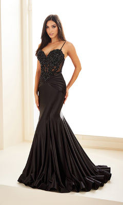 A woman in the Ellie Wilde EW36004 drop-waist mermaid prom dress, featuring a fitted black silhouette with a beaded bodice, thin straps, and flared satin skirt, stands indoors by a window.