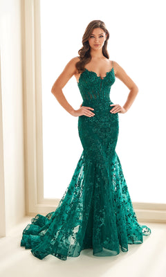 A woman stands near a window in the Ellie Wilde EW36008, a long, emerald green embroidered prom dress featuring a fitted lace-up bodice, thin straps, and a flared mermaid hem.