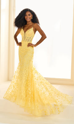 A woman stands in a bright room wearing the Ellie Wilde EW36008, a long embroidered prom dress with thin straps and a sheer, flared train.