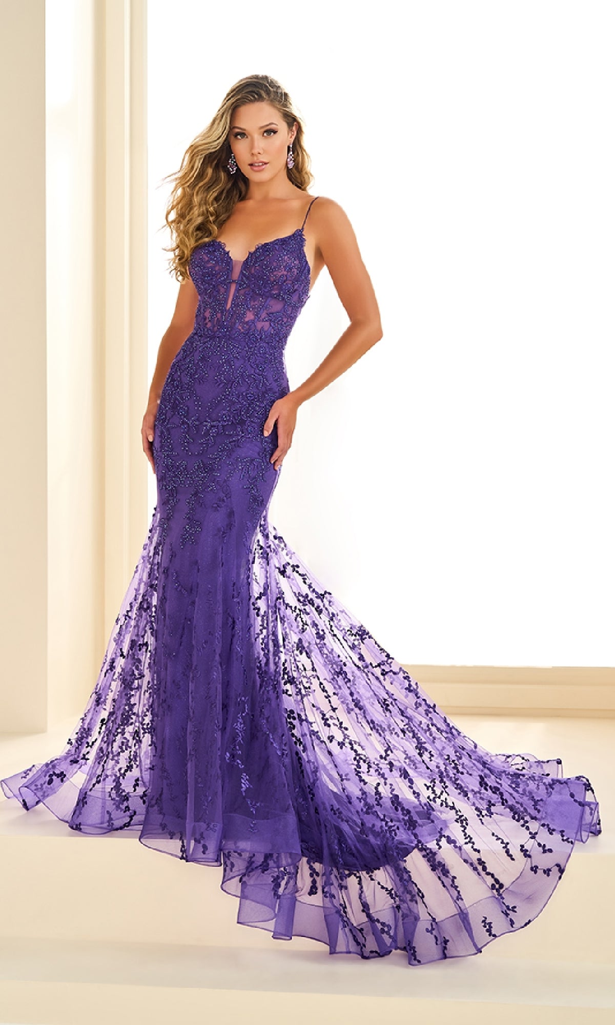 A woman stands indoors against a light background wearing the Ellie Wilde EW36008, a fitted, floor-length purple prom dress with lace embroidery and a flared sheer train.