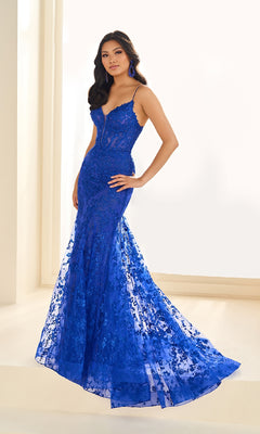 A woman in the Ellie Wilde EW36008, a fitted, floor-length royal blue embroidered prom dress with lace and thin straps, stands indoors against a light background.
