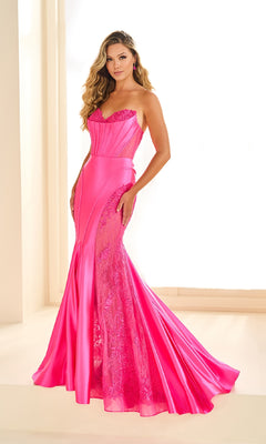 A woman poses indoors in the Ellie Wilde EW36010 long prom dress—a bright pink strapless mermaid gown featuring lace, satin details, and elegant sequin accents, with one hand on her hip.