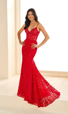 A woman poses indoors by a bright window in the Ellie Wilde EW36011, a form-fitting, floor-length red lace gown with thin straps.