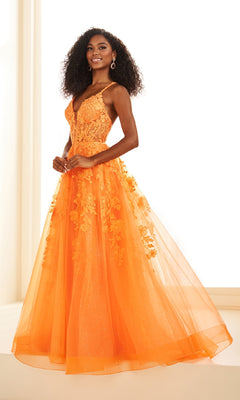 A woman poses indoors against a bright background, wearing the Ellie Wilde EW36015 long prom dress—a sleeveless, floor-length orange gown featuring lace embroidery and a tulle skirt.