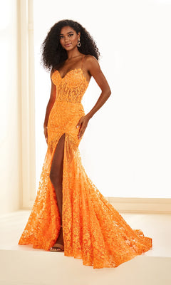 A woman poses indoors against a white background wearing the Ellie Wilde EW36016 Lace Long Prom Dress, featuring a bright orange hue, thigh-high slit, thin straps, and a fitted corset bodice.