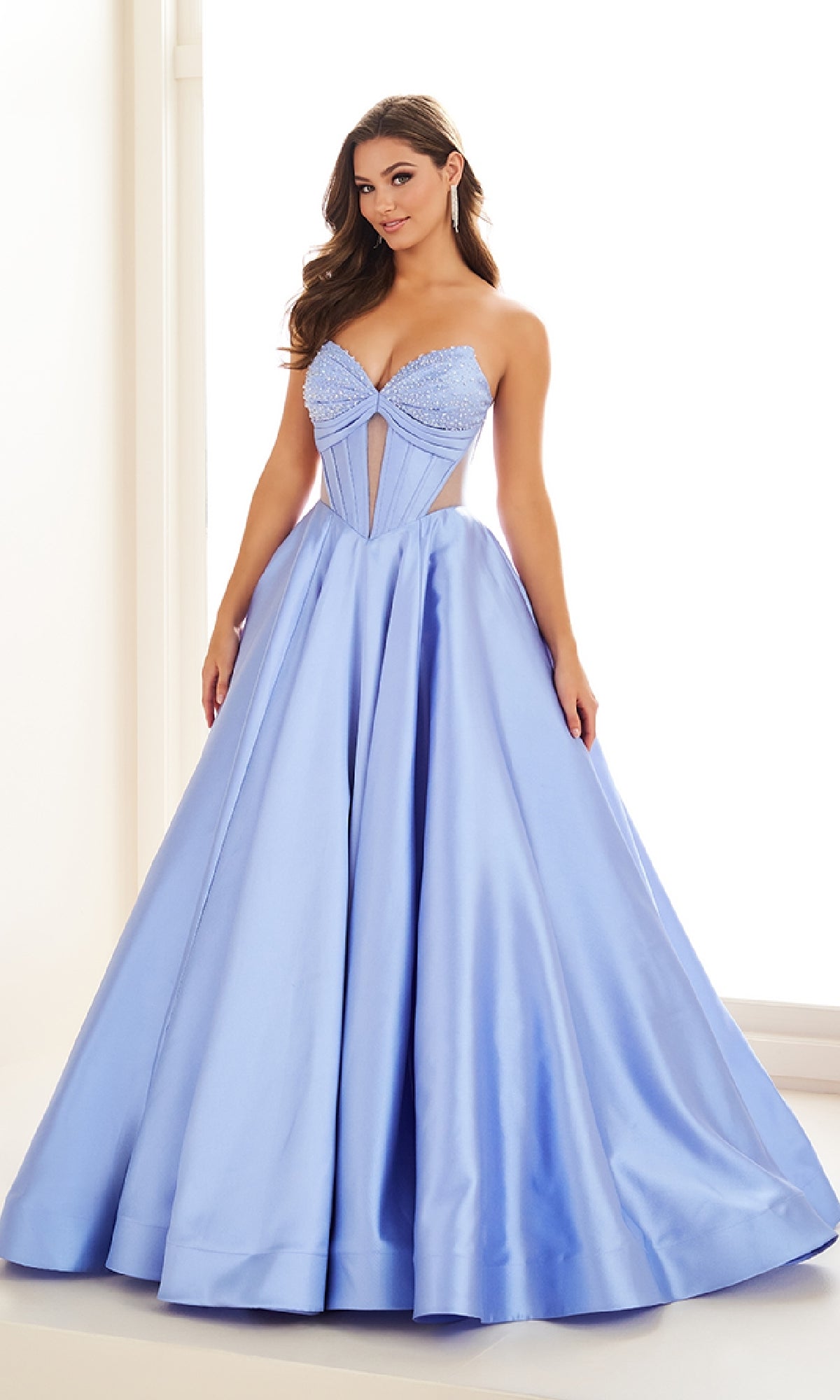 A woman models the Ellie Wilde EW36025, a strapless, floor-length light blue satin prom gown with a fitted corset bodice and full skirt, standing before a bright backdrop.
