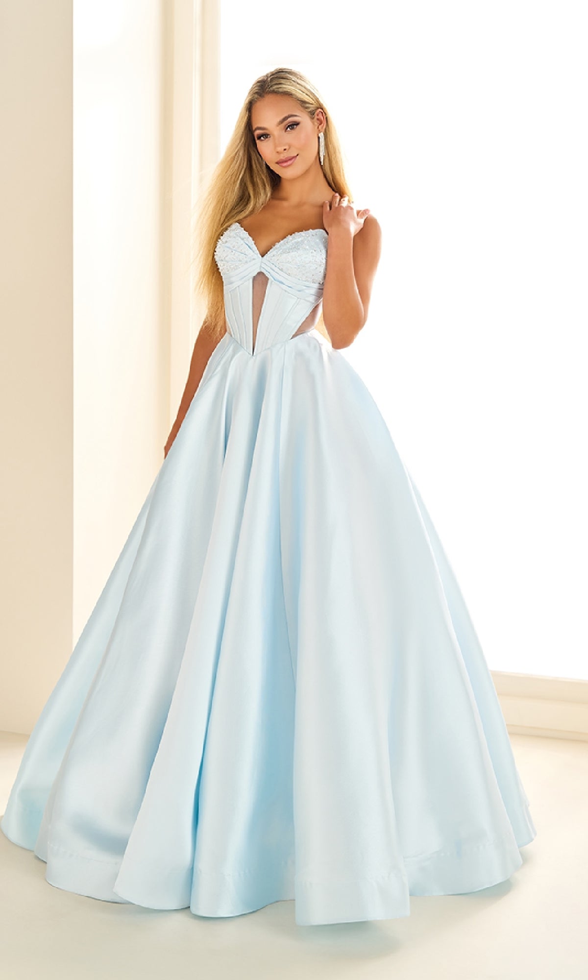 Woman in the Ellie Wilde EW36025 long prom dress, a light blue satin ball gown with a fitted corset bodice, full skirt, and lace-up open back, stands indoors by a window.