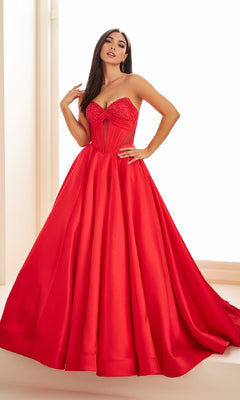 A woman stands indoors wearing the Ellie Wilde EW36025, a strapless, floor-length satin prom ball gown with a fitted corset bodice and full skirt.