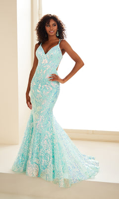 A woman poses indoors by a large window wearing the Ellie Wilde EW36034, a light blue, floor-length prom dress with floral sequins, thin straps, and a flared hem.