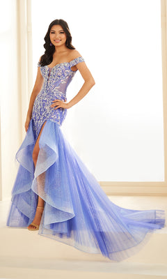 A woman stands indoors wearing the Ellie Wilde EW36036, a lavender off-the-shoulder long prom dress with a corset bodice, silver embroidery, layered tulle skirt, and glittery mermaid-style details.