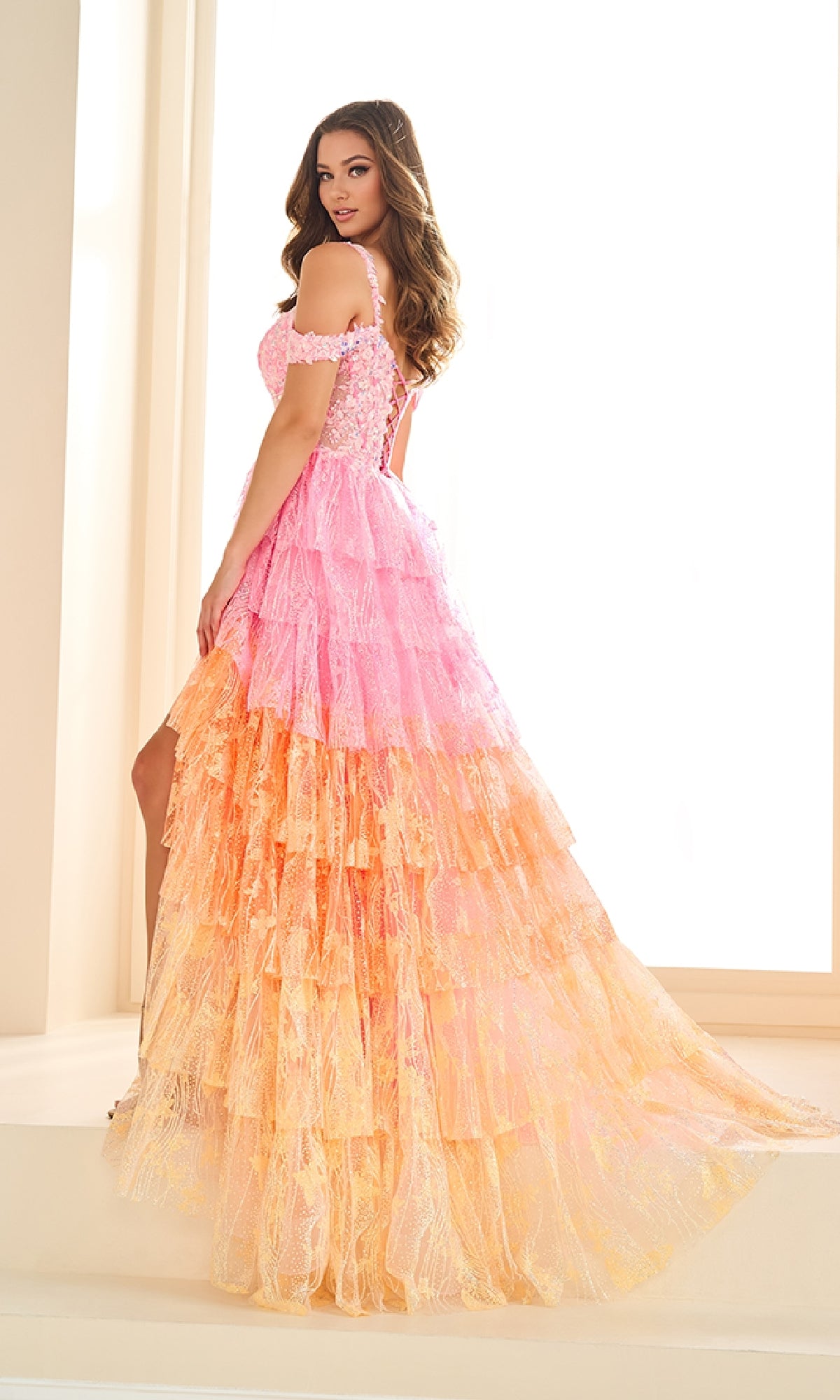 A woman stands sideways on a white staircase, looking toward the camera in a brightly lit room, wearing the Long Beaded-Lace Prom Dress: Ellie Wilde EW36037.