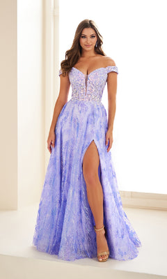 A woman stands indoors against a light background wearing the Ellie Wilde EW36039 Glow-in-the-Dark Prom Dress, featuring an off-the-shoulder beaded bodice, front slit, and floor-length lavender skirt.