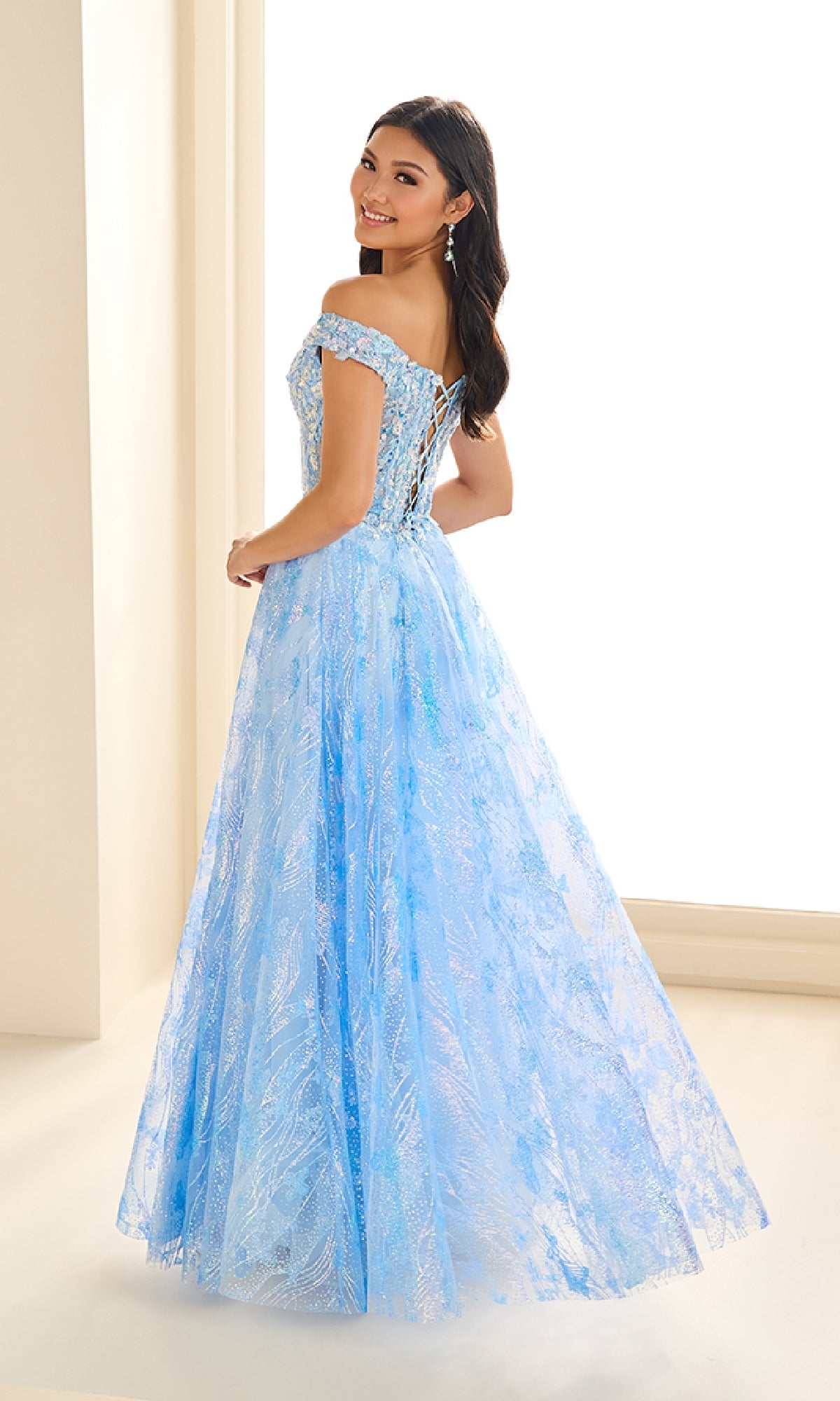 A woman smiles indoors, looking over her shoulder in the Ellie Wilde EW36039 Glow-in-the-Dark Prom Dress—a light blue, off-the-shoulder gown with a corset bodice, lace-up back, and full skirt.