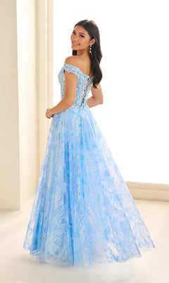 A woman smiles indoors, looking over her shoulder in the Ellie Wilde EW36039 Glow-in-the-Dark Prom Dress—a light blue, off-the-shoulder gown with a corset bodice, lace-up back, and full skirt.