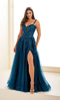 A woman poses indoors near a window wearing the Long Prom Dress: Ellie Wilde EW36046, a deep teal, floor-length gown with sequin lace applique, sweetheart neckline, and high slit.