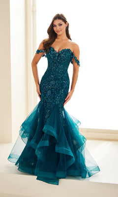A woman stands indoors wearing the Ellie Wilde EW36047, a teal off-the-shoulder mermaid prom dress with sequins, glitter tulle ruffles, and floral appliqué detailing.