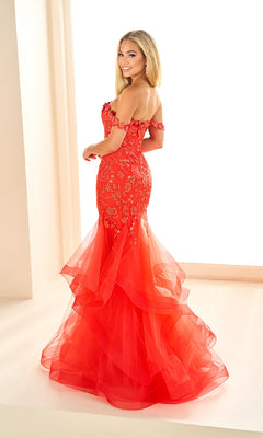 A woman stands indoors wearing the Ellie Wilde EW36047 long prom dress, an off-shoulder fitted red mermaid style with glitter tulle and floral details, looking over her shoulder and smiling.