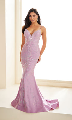 A woman poses indoors by a large window wearing the Ellie Wilde EW36048—a light purple, fitted long prom dress with sequins, thin straps, and a mermaid silhouette.
