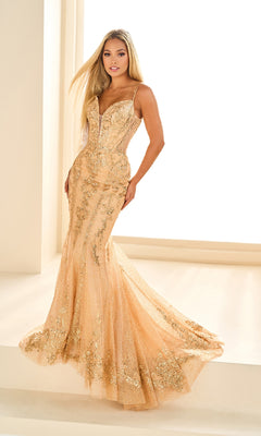 A woman stands in a brightly lit room wearing the Ellie Wilde EW36056 long gold fitted mermaid prom dress, featuring thin straps and floral embellishments.