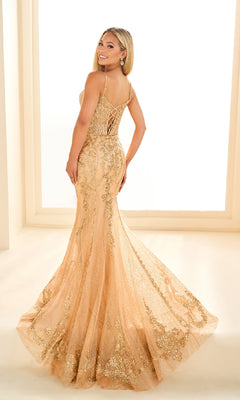 A woman smiles over her shoulder in the Ellie Wilde EW36056 gold, floor-length mermaid prom dress with lace applique details and a lace-up back.